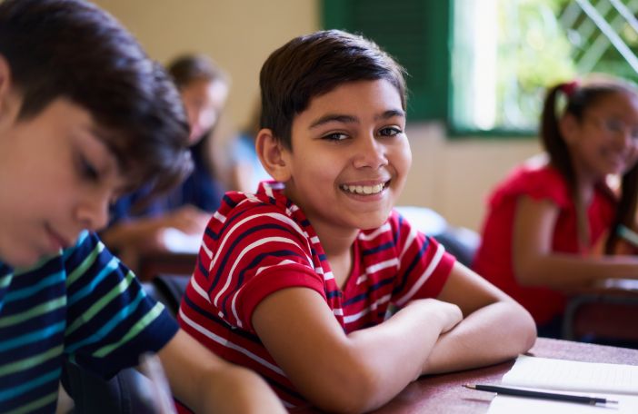 A young boy in school smiling at the camera from his desk.